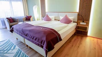 Una cama grande con cubrecama morado, una mesita de noche con lámpara, y una silla en una habitación de hotel con pisos de madera y un ventanal amplio.