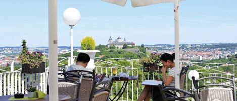 Una terraza con sillas y mesas de mimbre, con vistas a un paisaje urbano y un edificio parecido a un castillo.