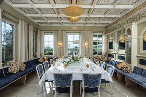 Uma sala de jantar formal com uma longa mesa posta para uma refeição, rodeada por assentos estofados em azul.