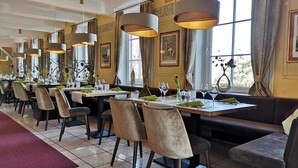 Un restaurante con varias mesas y sillas, ventanas amplias con cortinas, y lámparas colgantes.