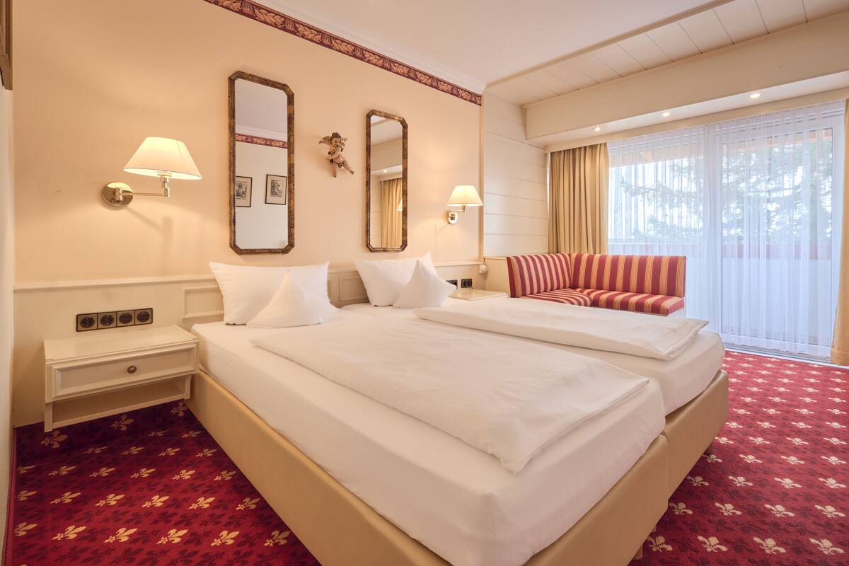 A hotel room with two beds, a red carpet, a large window with curtains, and wall-mounted lamps.