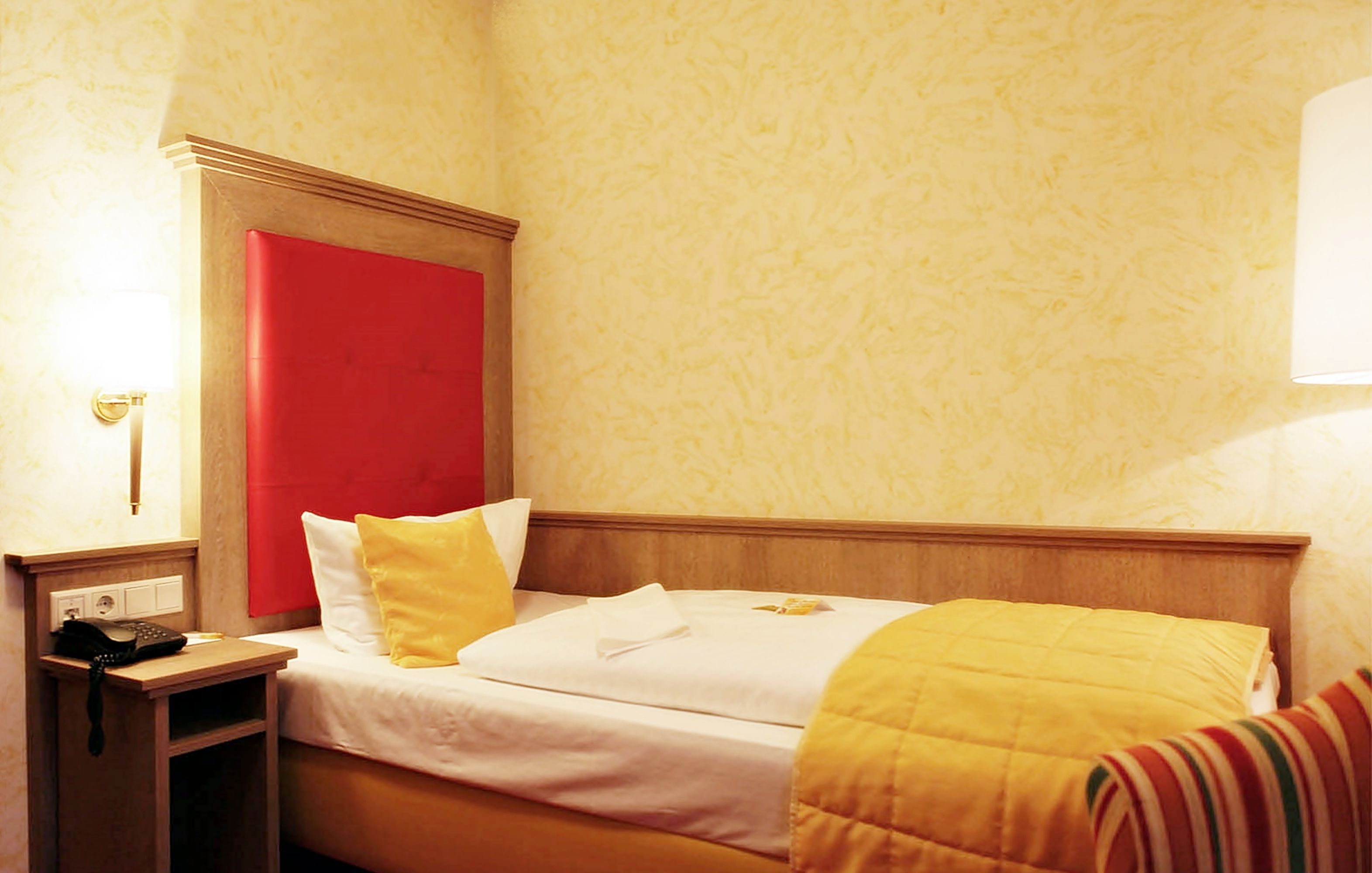 A hotel room with a bed, a nightstand, a lamp, and a red accent wall.