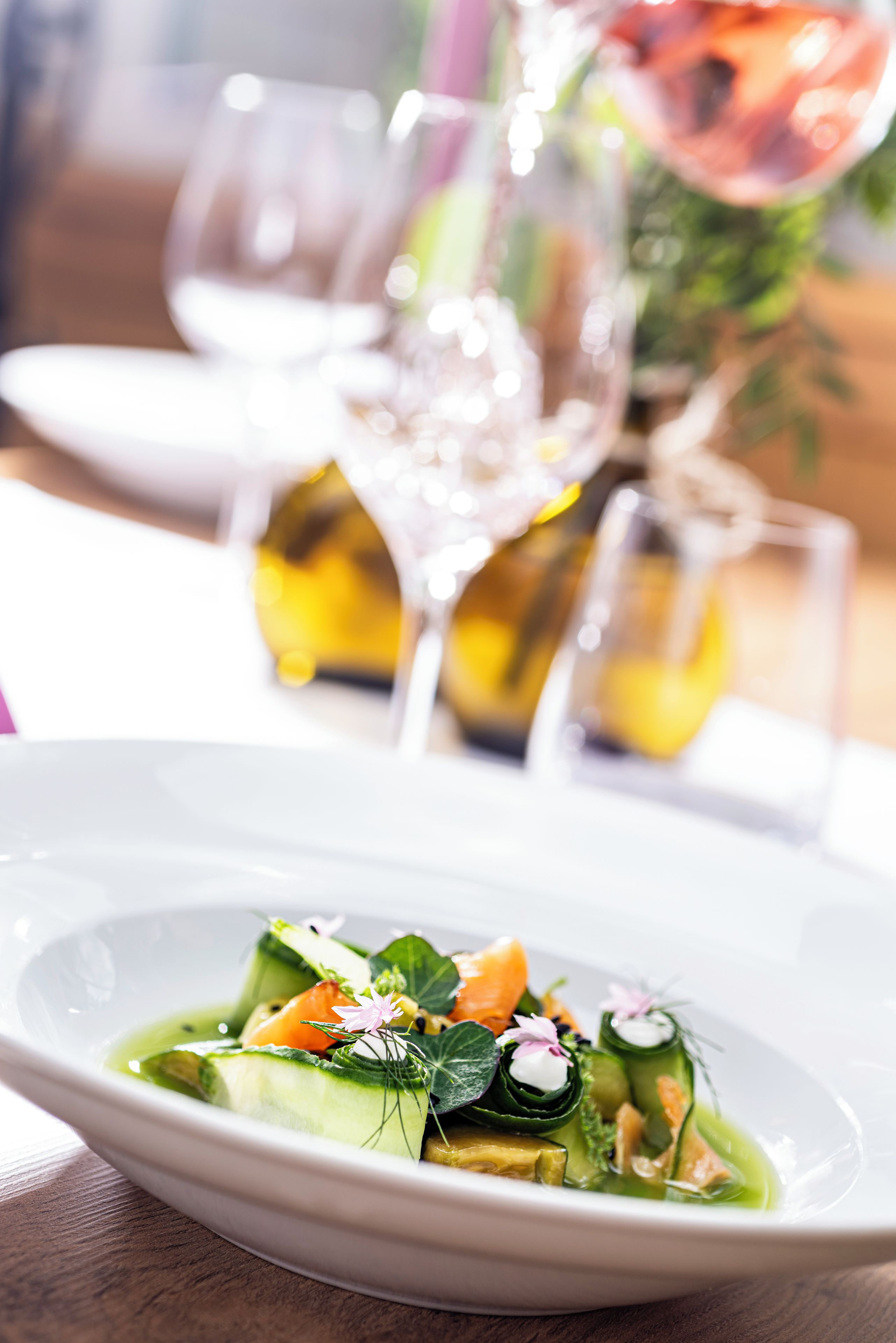 Ein Teller mit Essen, wobei das Gericht im Mittelpunkt steht, begleitet von einem Weinglas und einer Vase mit Blumen im Hintergrund.