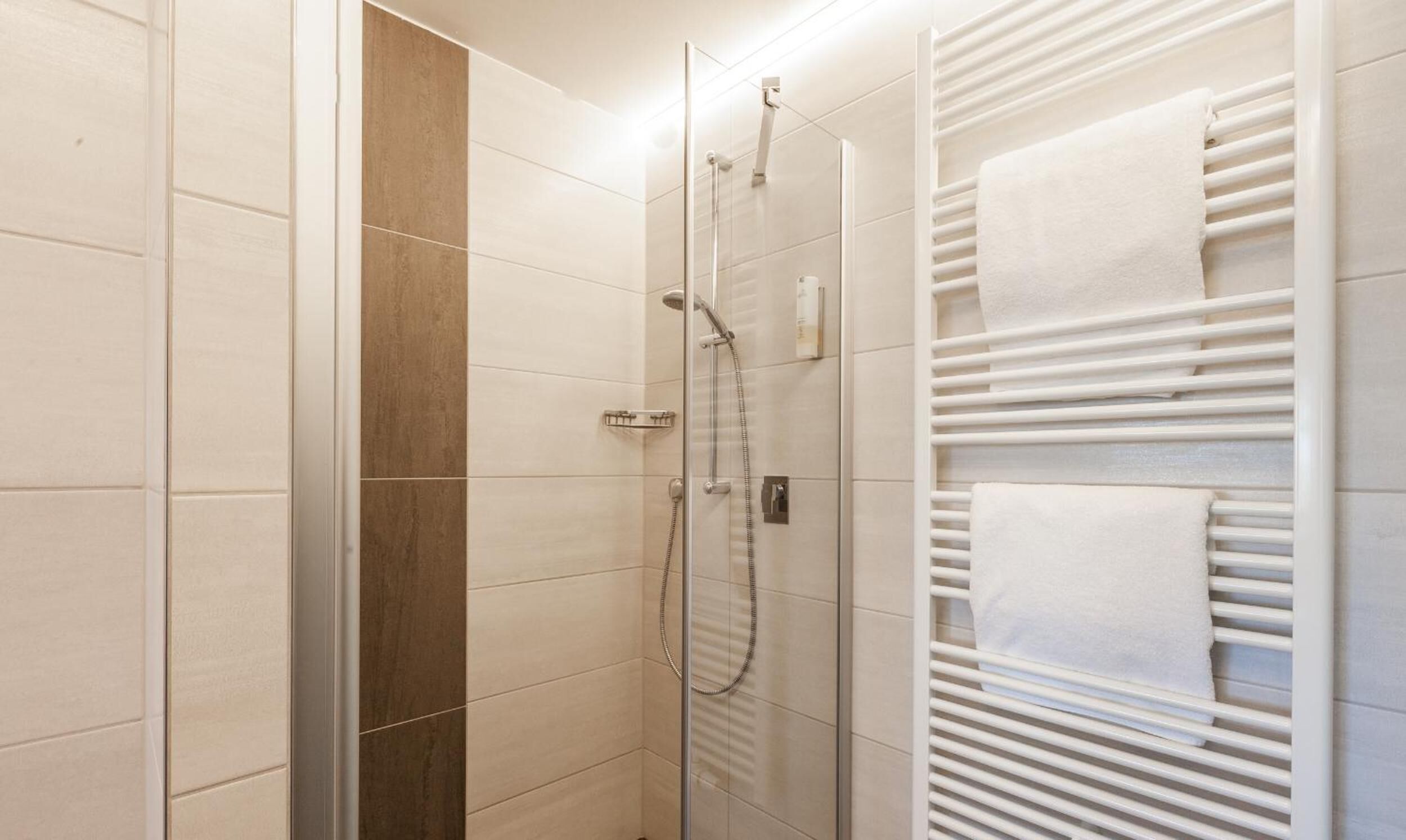 Un baño moderno con cabina de ducha de vidrio, toallero blanco y pared con azulejos color beige.