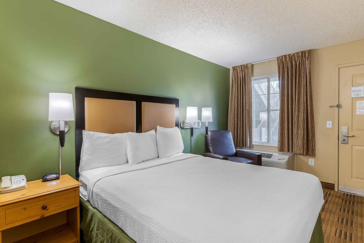 A hotel room with a large bed, two bedside lamps, a nightstand with a phone, a chair, and a window with curtains.