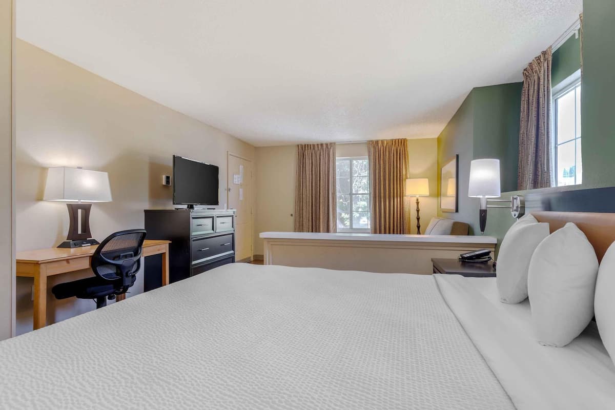 A hotel room with a large bed, a desk with a chair, a television, a window with curtains, and a nightstand with a lamp.