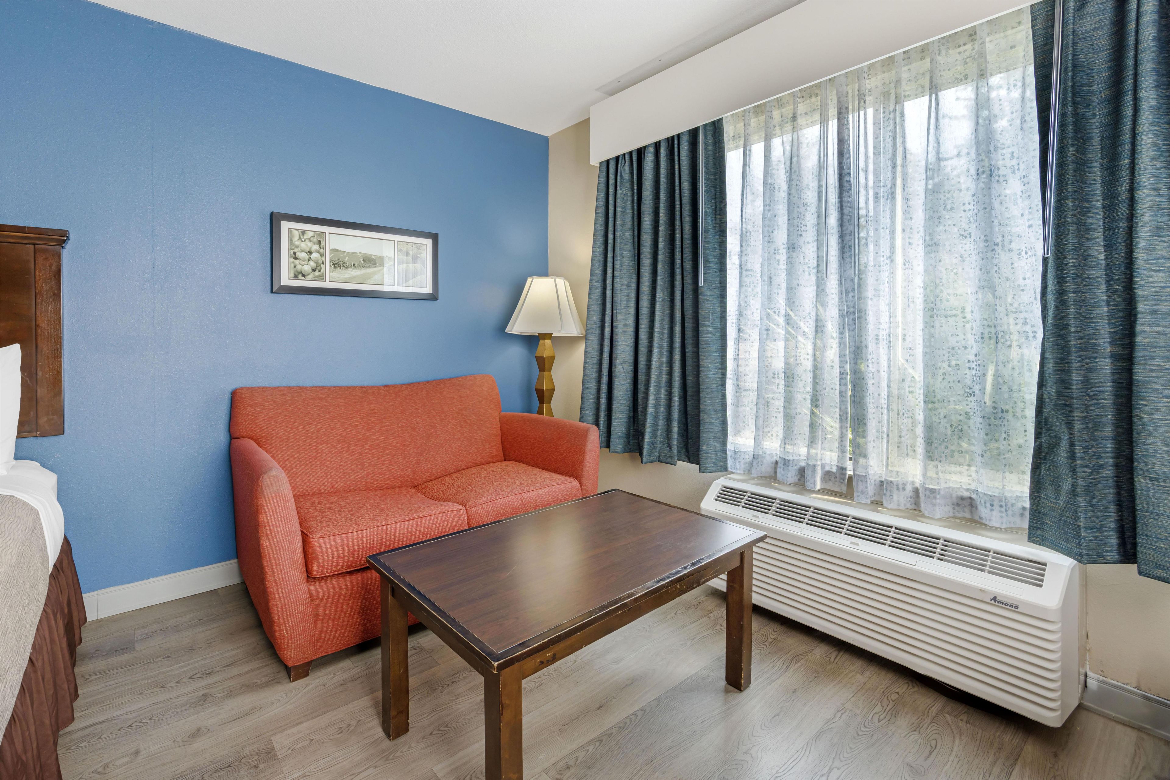 A hotel room with a blue wall, a red sofa, a wooden table, a lamp, and a window with curtains.