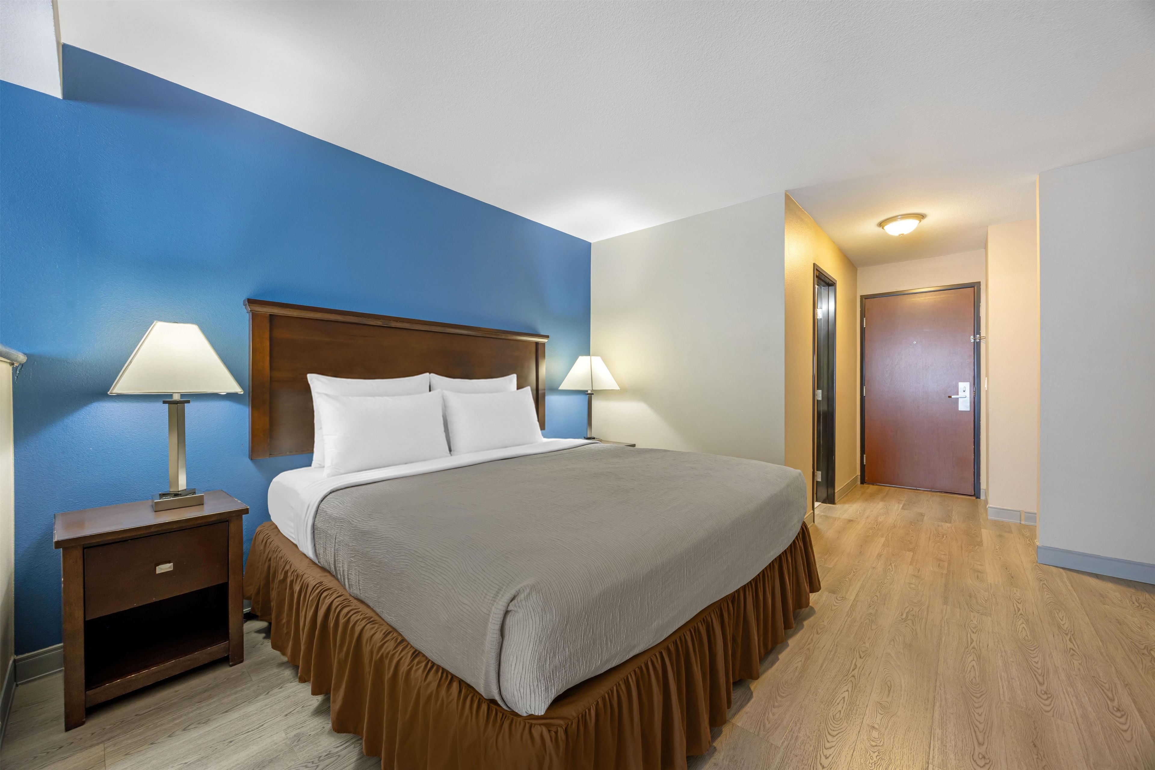 Ein Hotelzimmer mit einem großen Bett, zwei Nachttischlampen, einem Holznachttisch und einer Tür zu einem weiteren Raum.