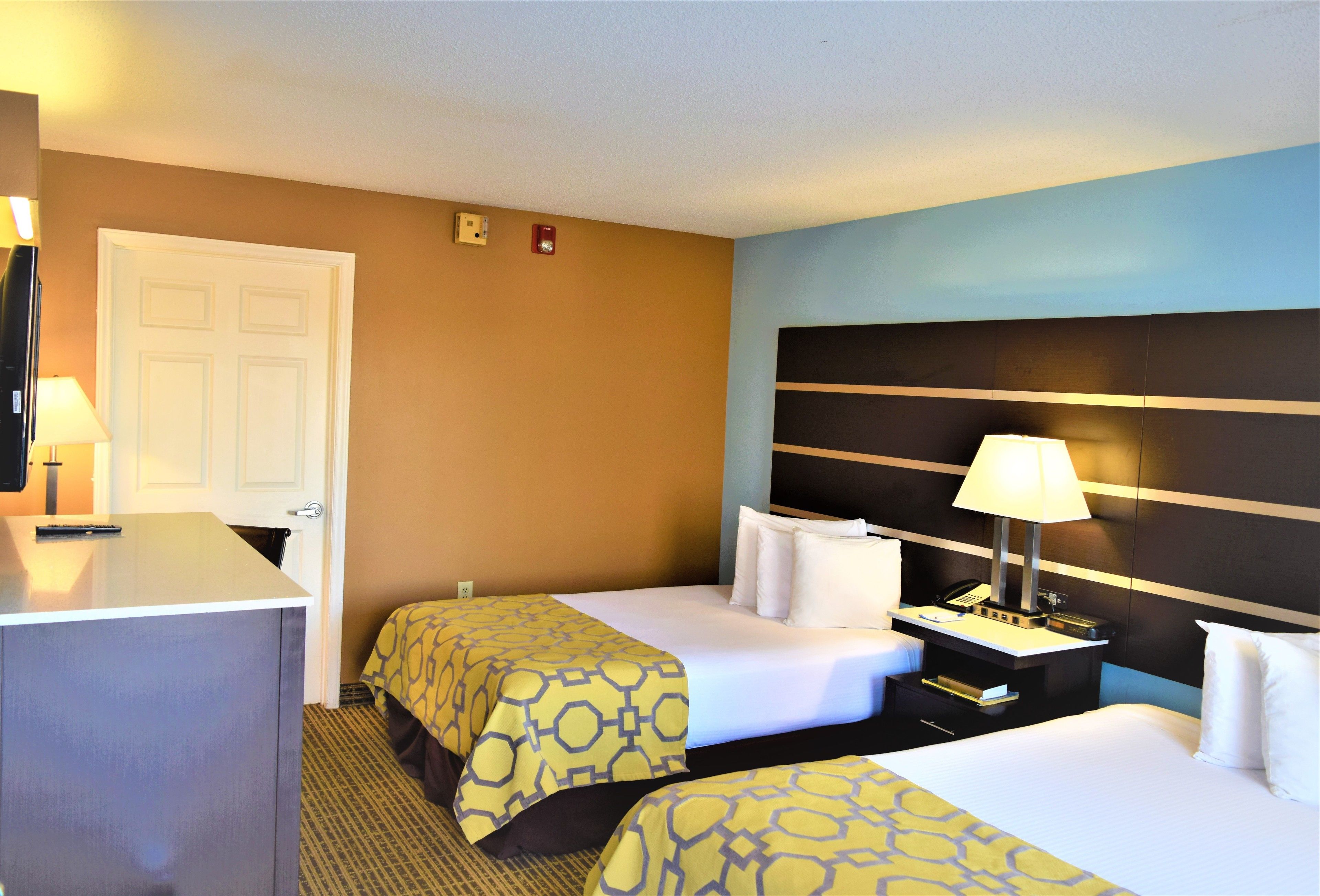 A hotel room with two beds, a desk, and a nightstand with a lamp.