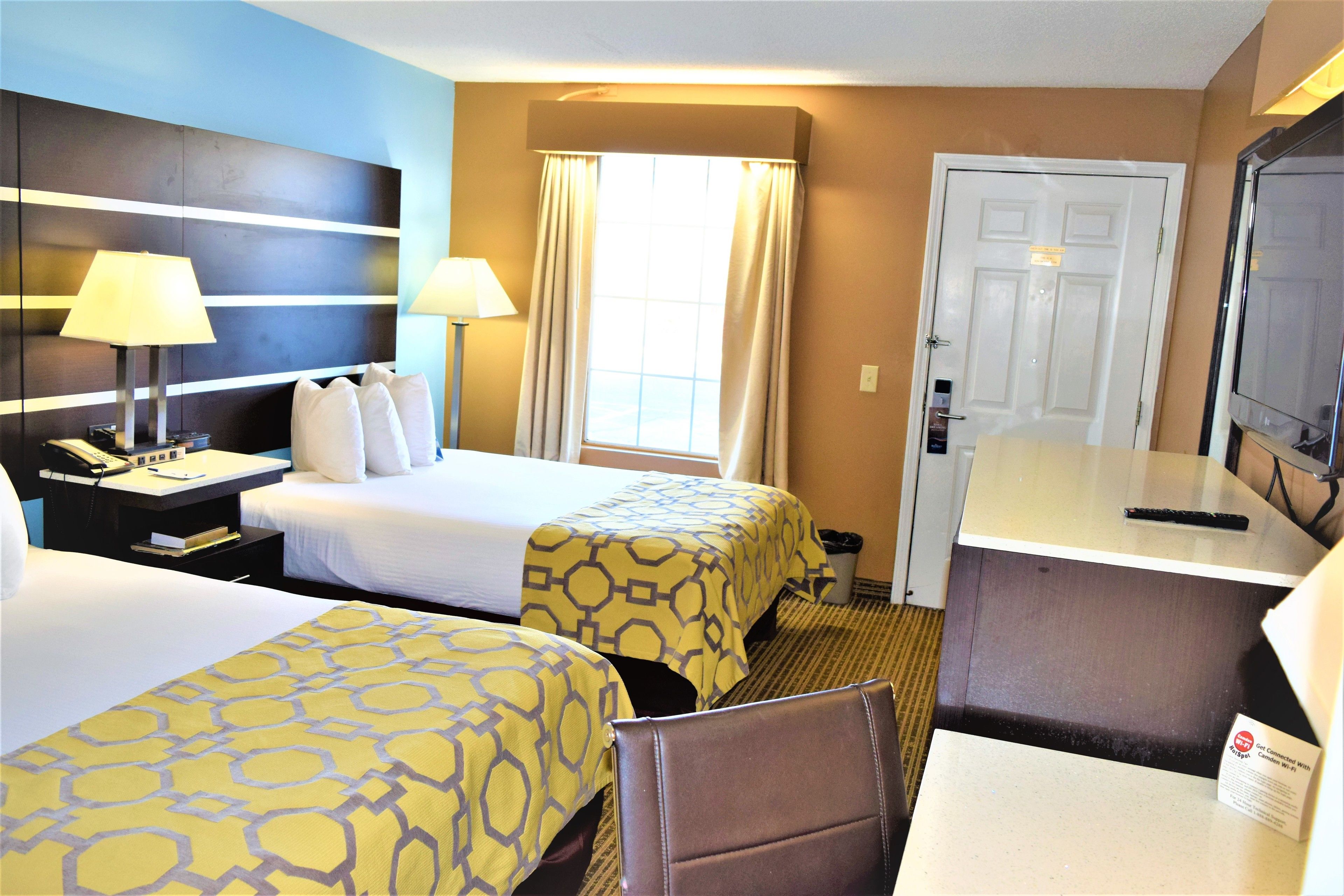 Quarto de hotel com duas camas, uma escrivaninha, uma cadeira, uma televisão e uma janela com cortinas.