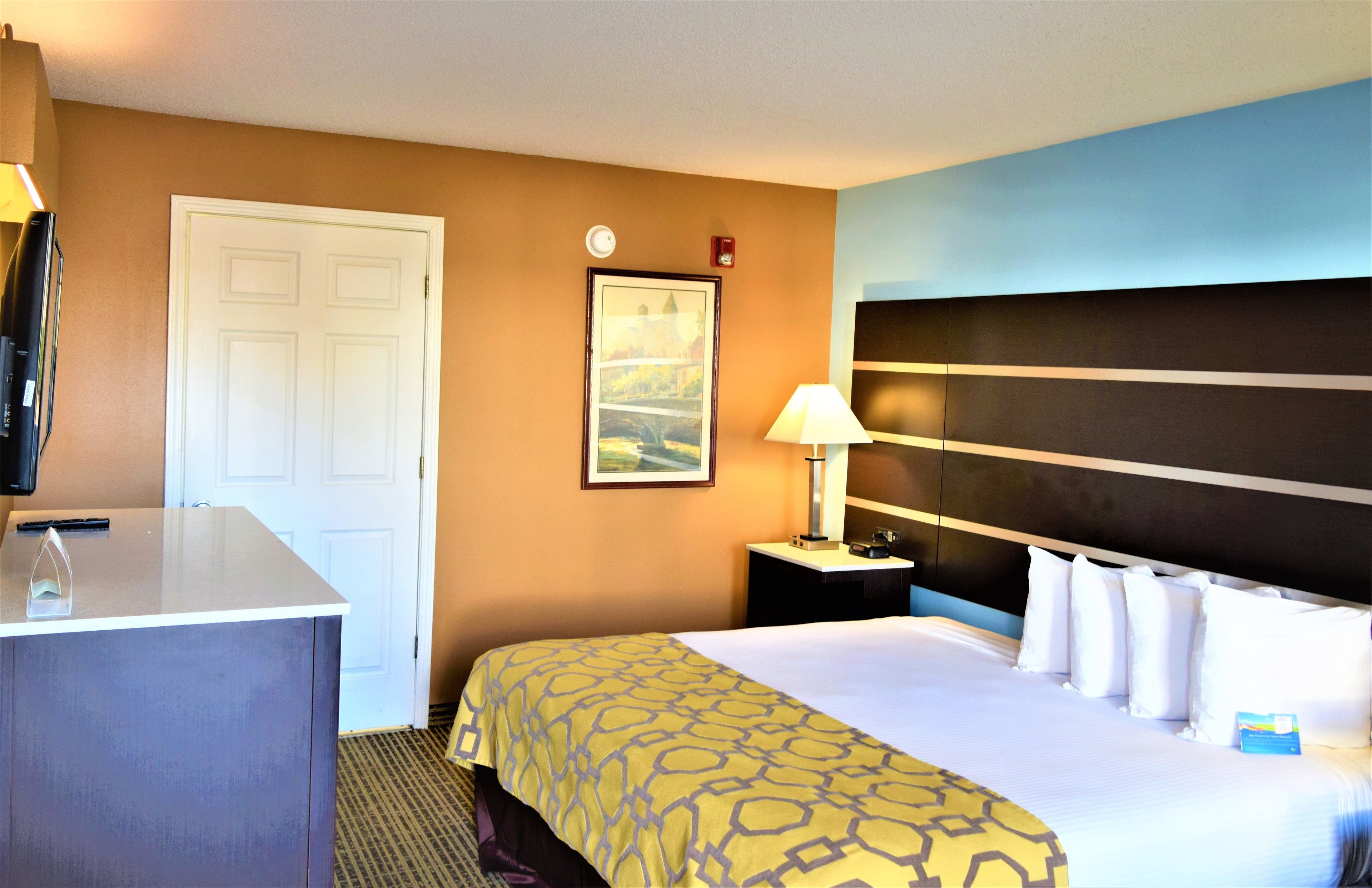 A hotel room with a bed, a desk, a lamp, a clock, and a framed picture on the wall.