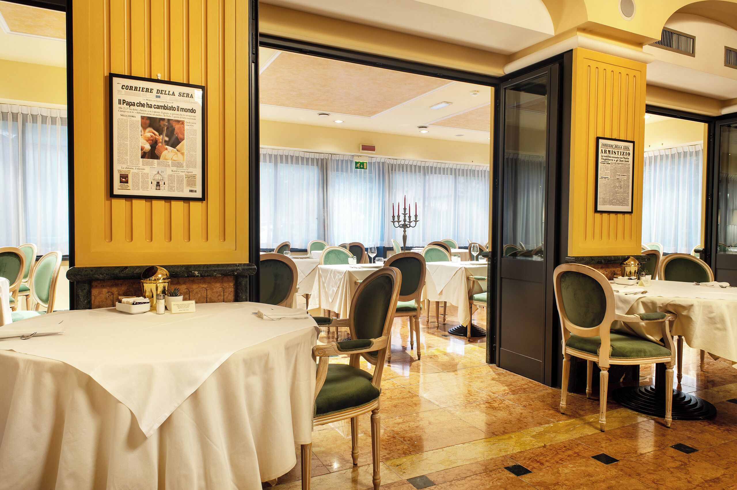A dining area with tables set for service, chairs with green upholstery, and a chandelier.
