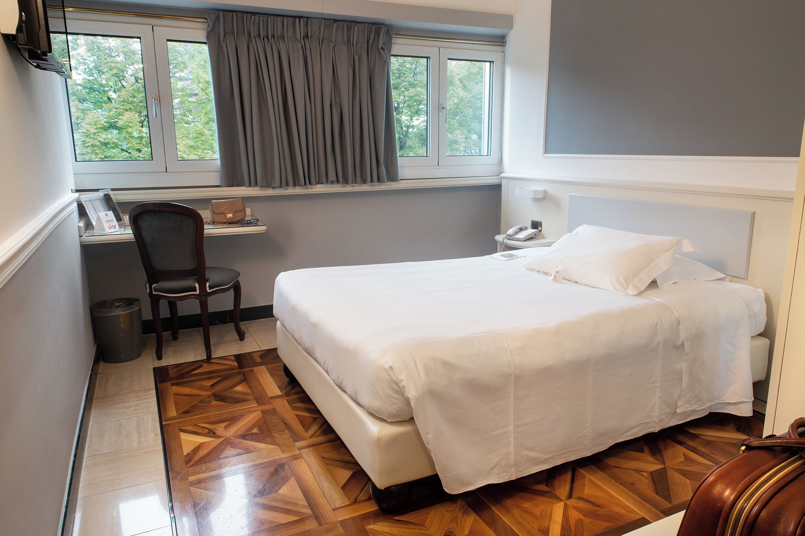 A hotel room with a bed, a desk with a chair, a telephone, and a window with curtains.