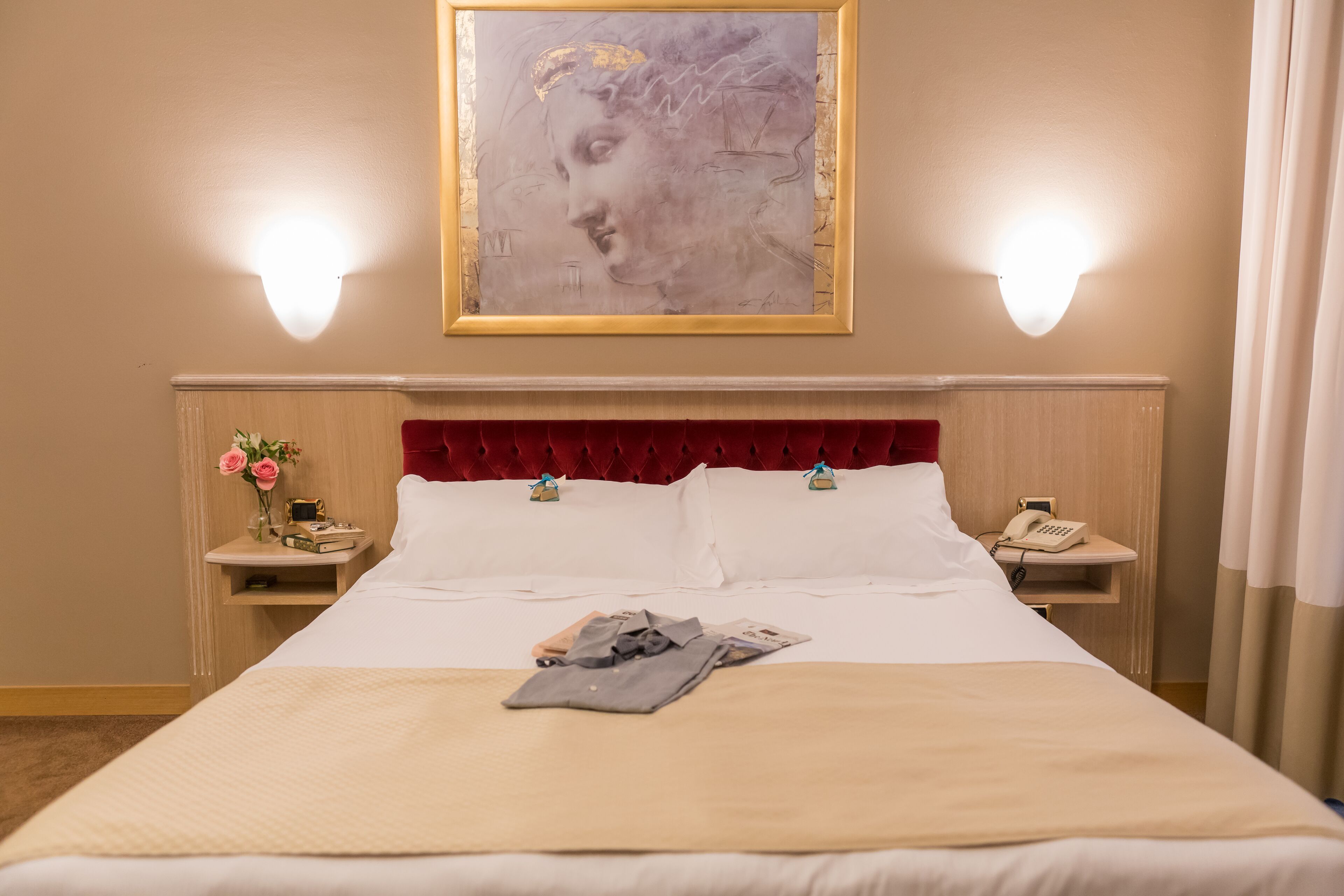 Habitación de hotel con una cama grande, dos mesitas de noche, un cabecero rojo, un cuadro sobre la cama y un teléfono en una de las mesitas.