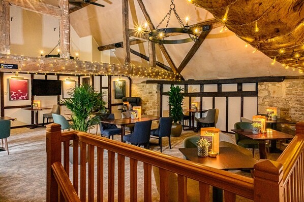 A cozy restaurant with wooden beams, stone walls, and string lights. Tables are set with chairs and candles.