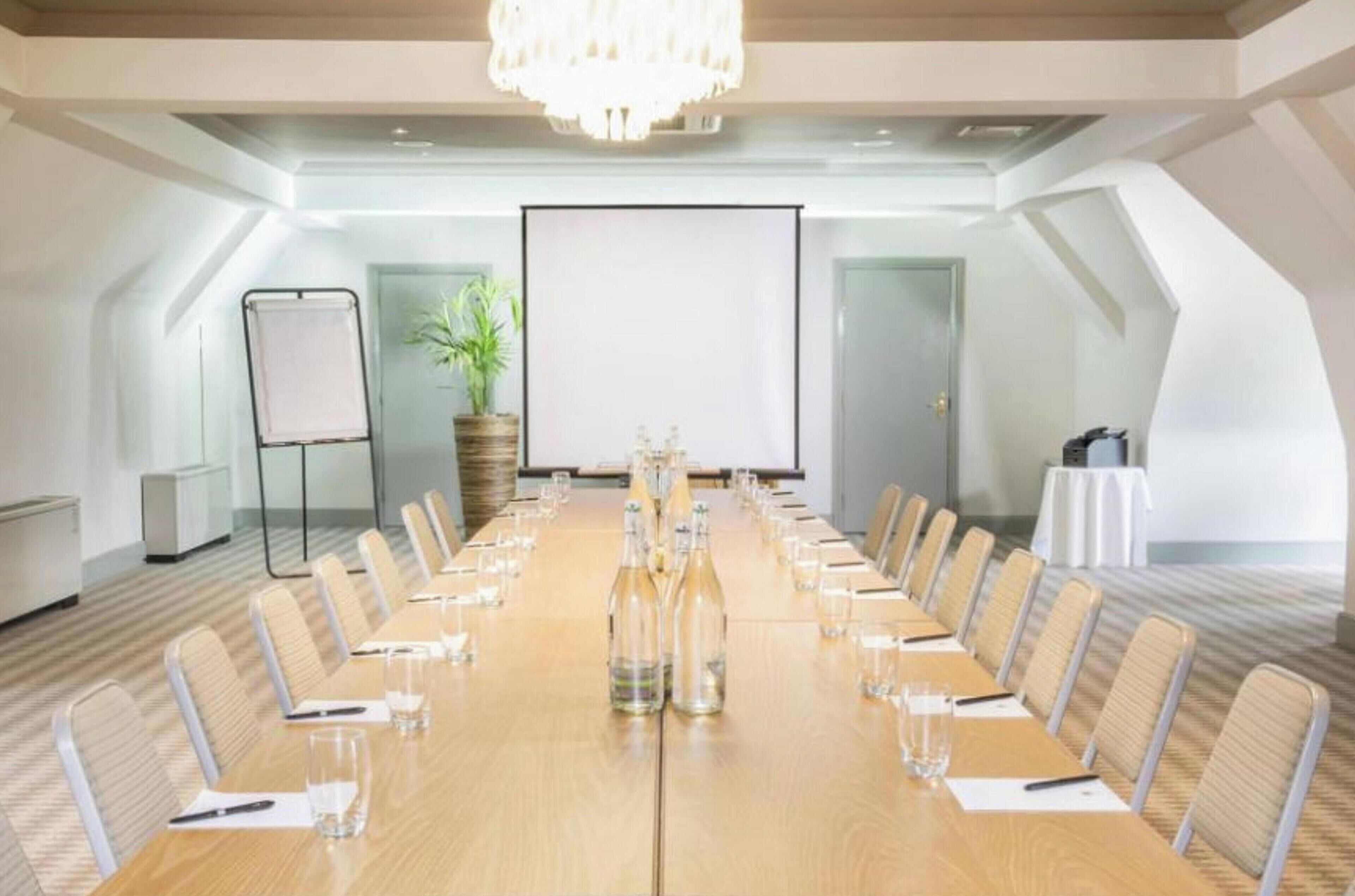 A conference room with a long table, chairs, a projector screen, and a potted plant.
