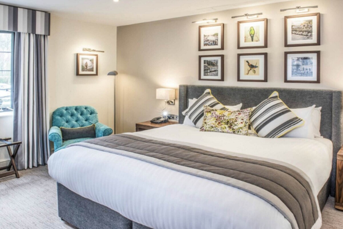 A hotel room with a large bed, a tufted armchair, a bedside table with a lamp, and framed pictures on the wall.
