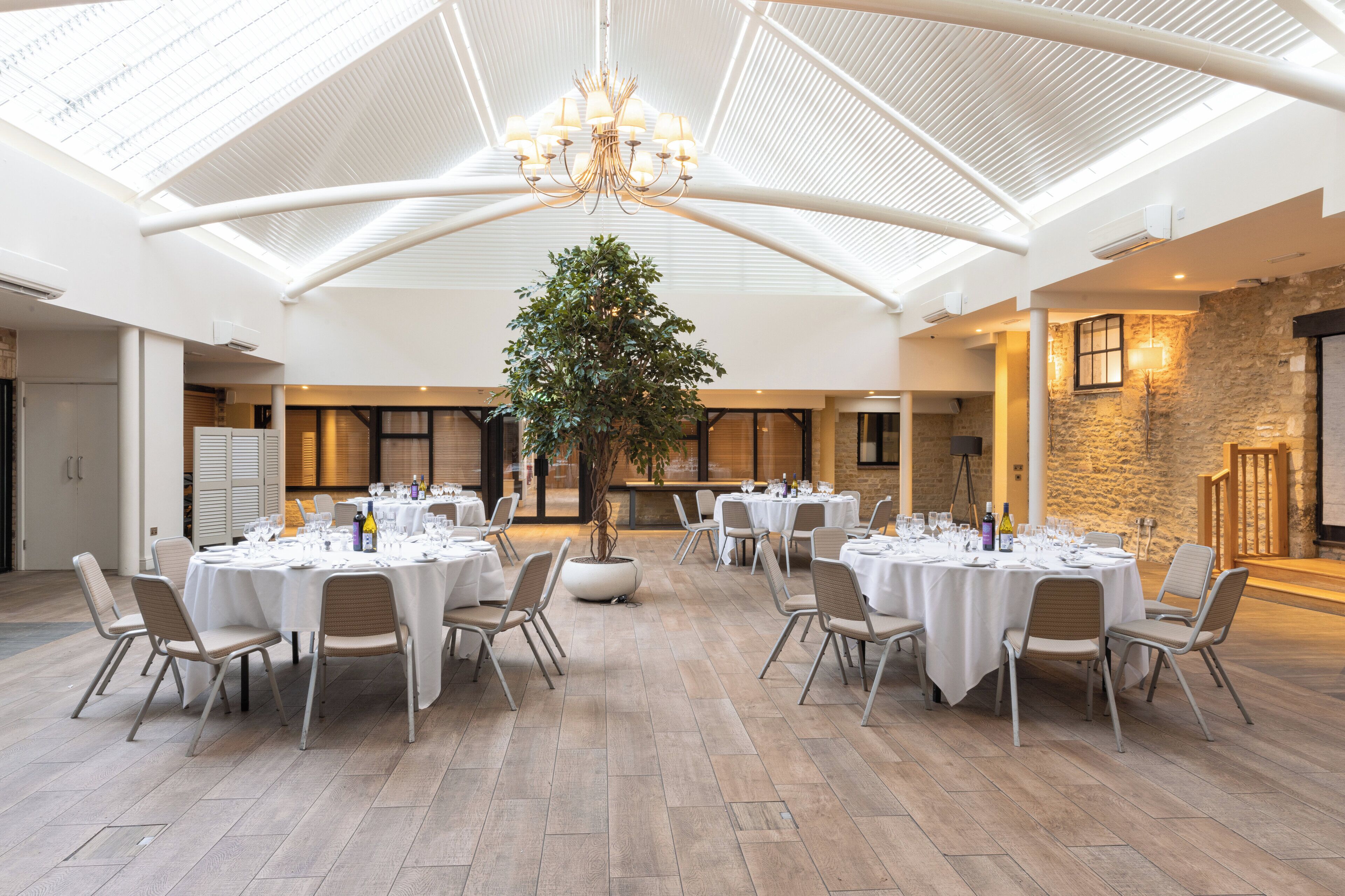 A spacious indoor dining area with round tables and chairs, a large tree, and a high ceiling with a skylight.