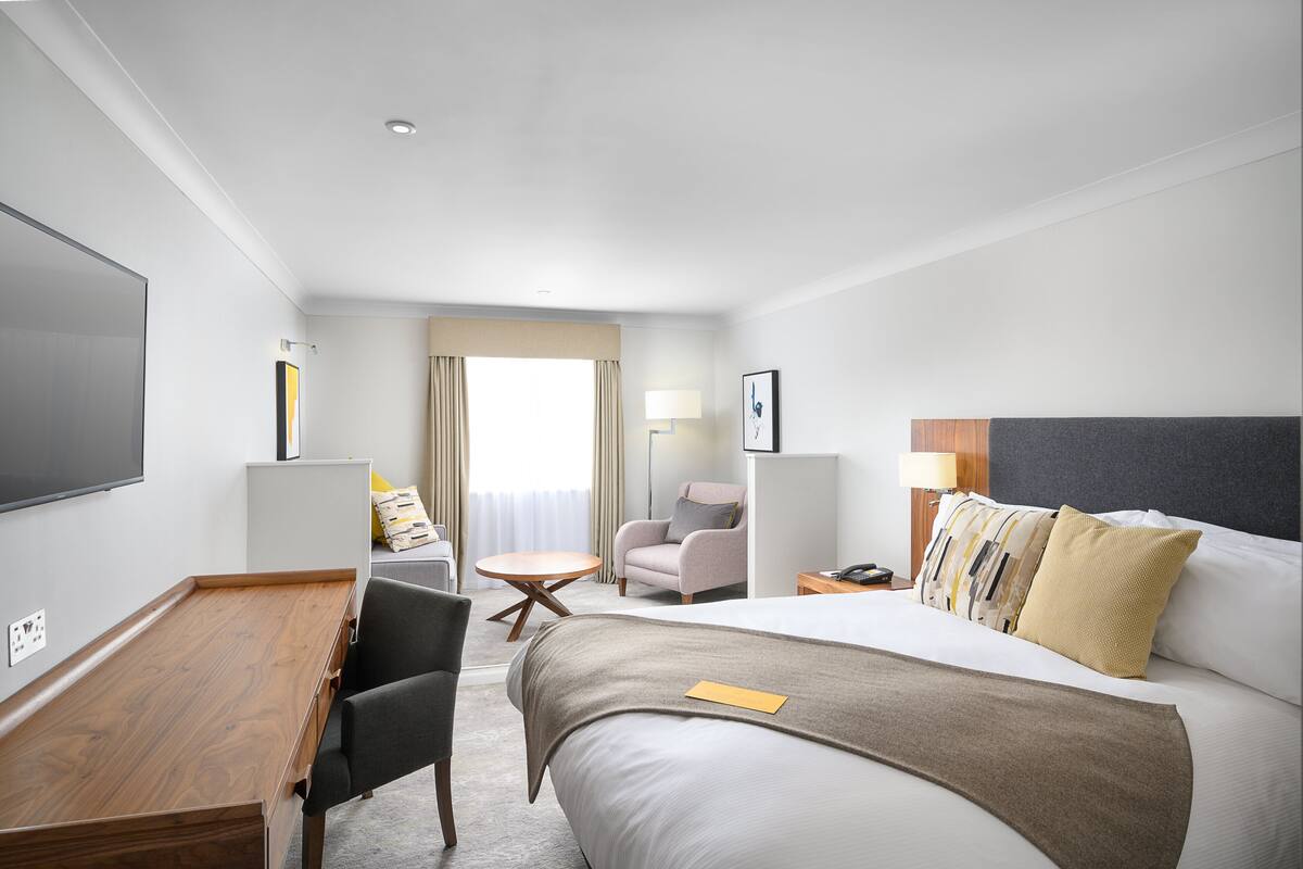 A hotel room with a bed, a desk, a chair, a small table, and a TV.
