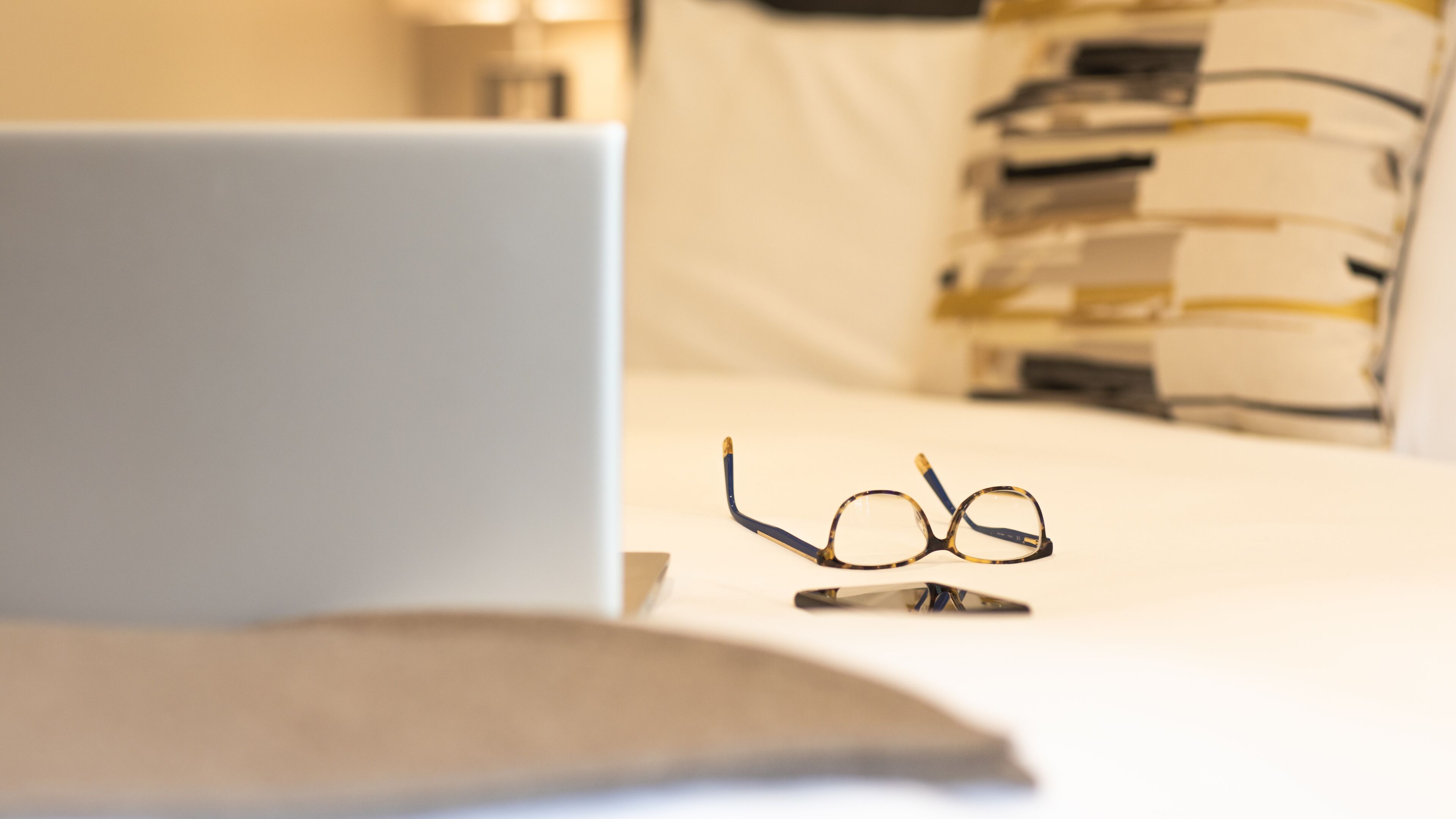 A laptop on a bed with a pair of glasses and a stack of books in the background.