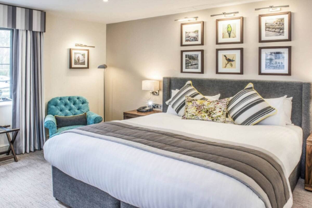 A hotel room with a large bed, a tufted armchair, a bedside table with a lamp, and framed pictures on the wall.