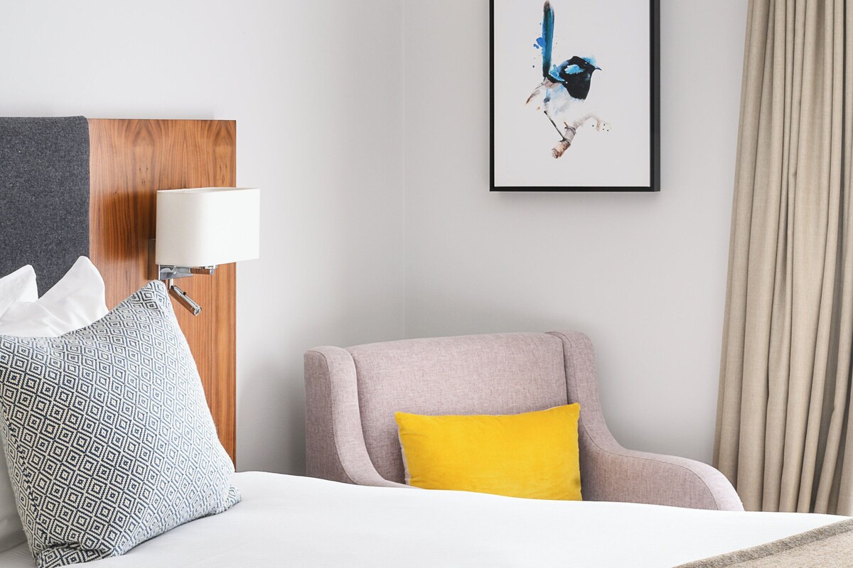 A hotel room with a bed, a chair, a lamp, a framed artwork, and a curtain.