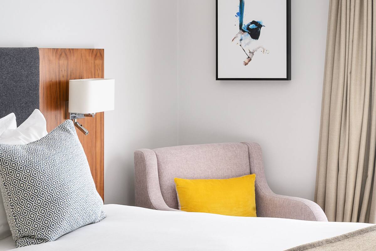 A hotel room with a bed, a chair, a lamp, a framed artwork, and a curtain.