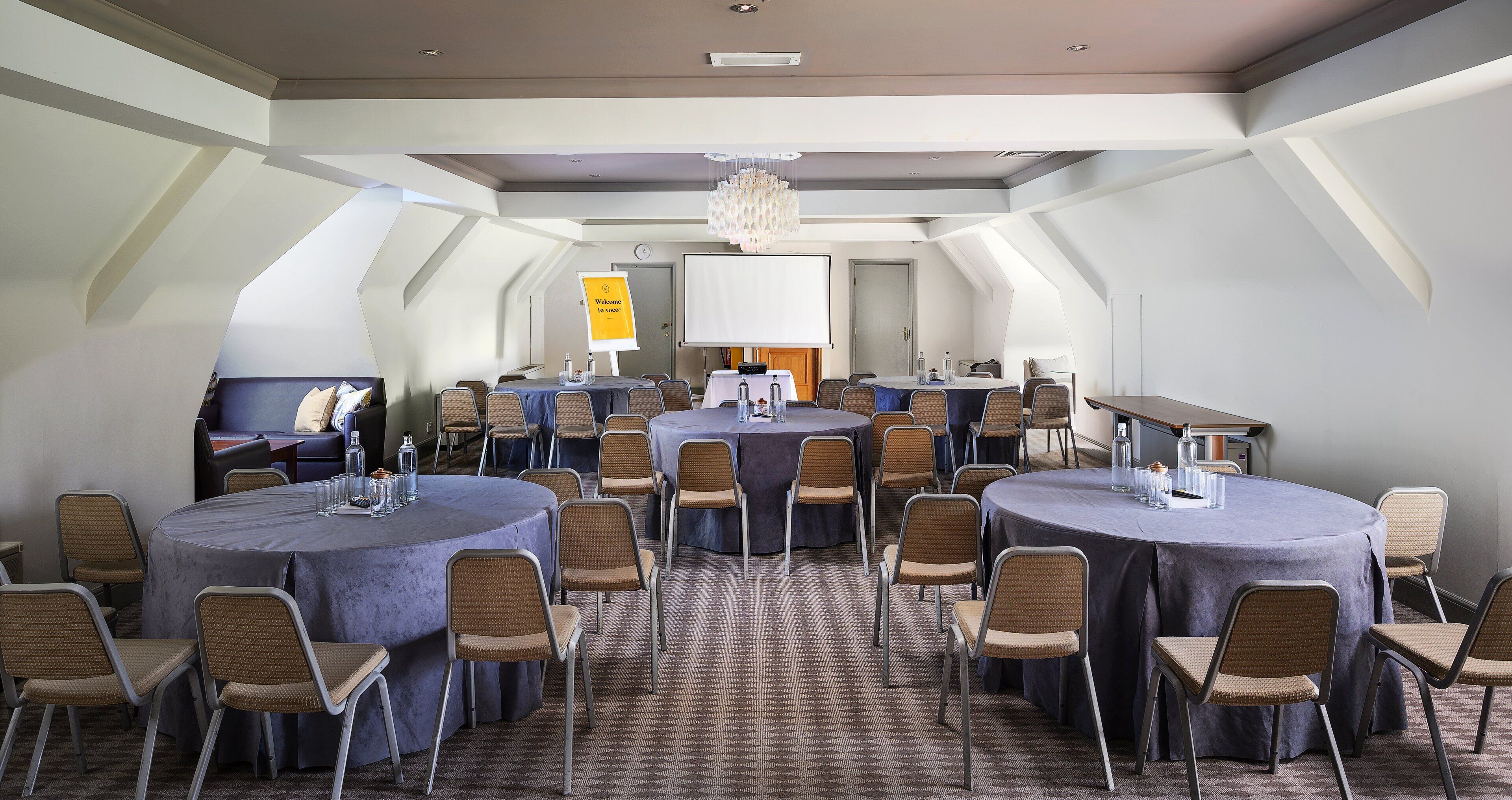 A conference room with multiple tables and chairs, a projector screen, and a welcome sign.