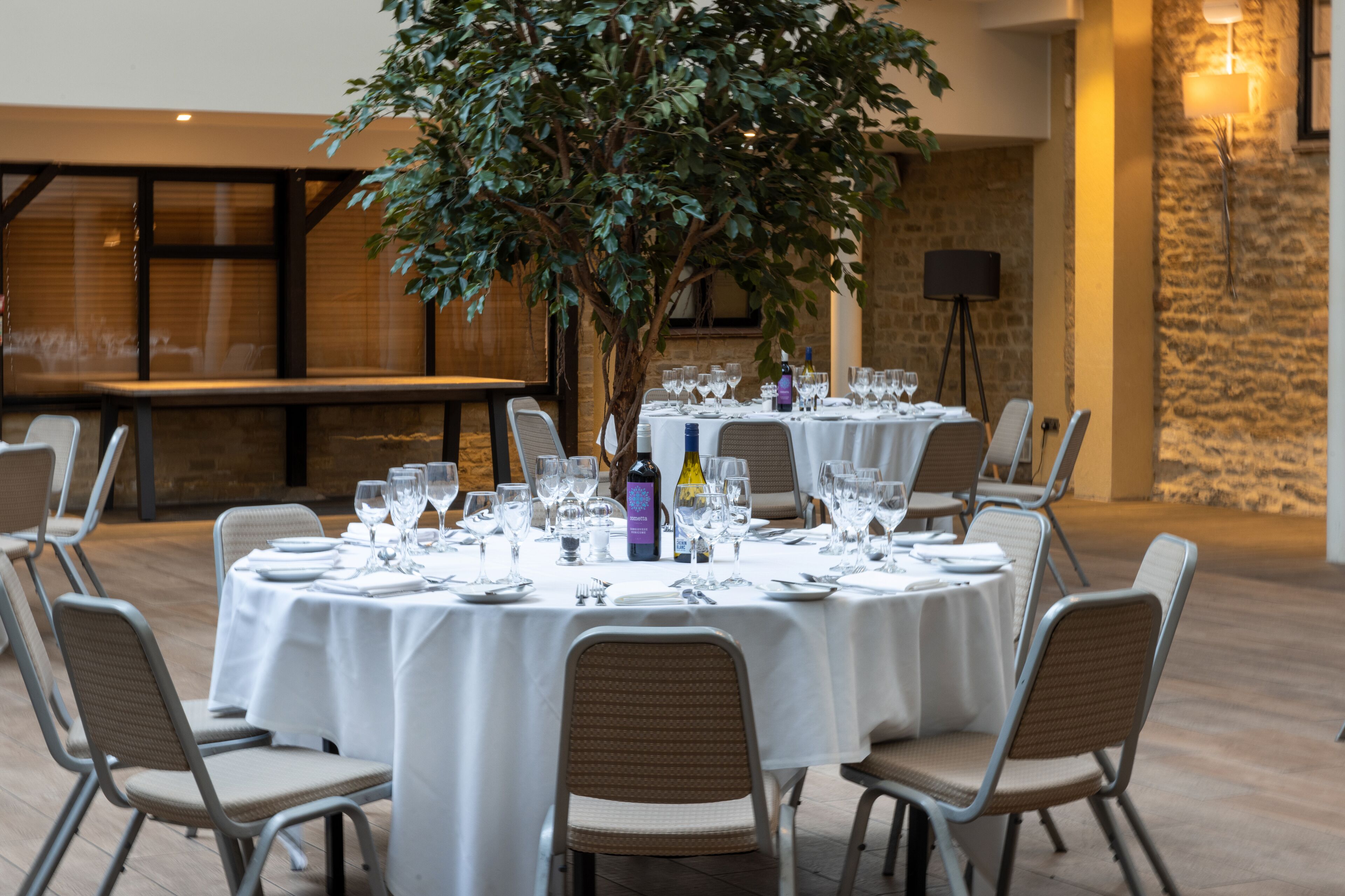 A round dining table set with white tablecloths, surrounded by chairs, in a well-lit indoor area with a large tree and stone walls.