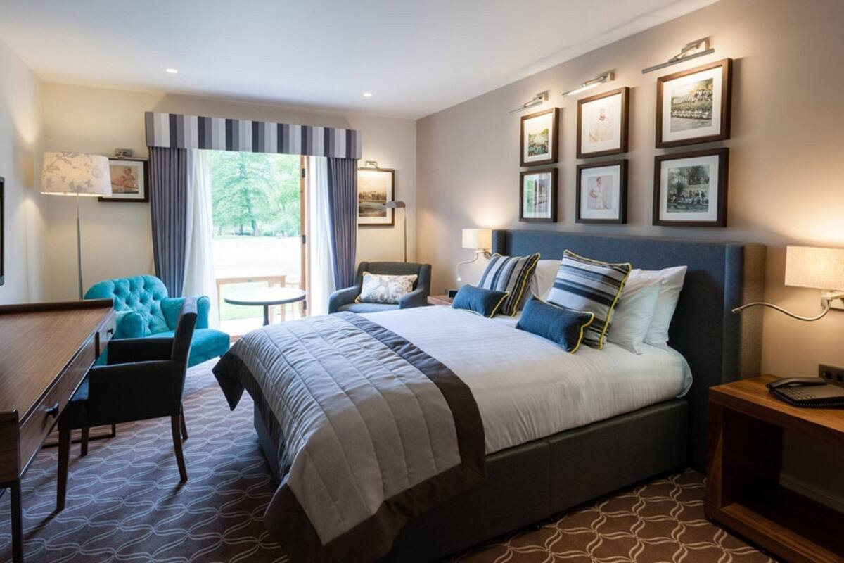 A modern hotel room with a large bed, a desk, a blue armchair, and a view of the outdoors.