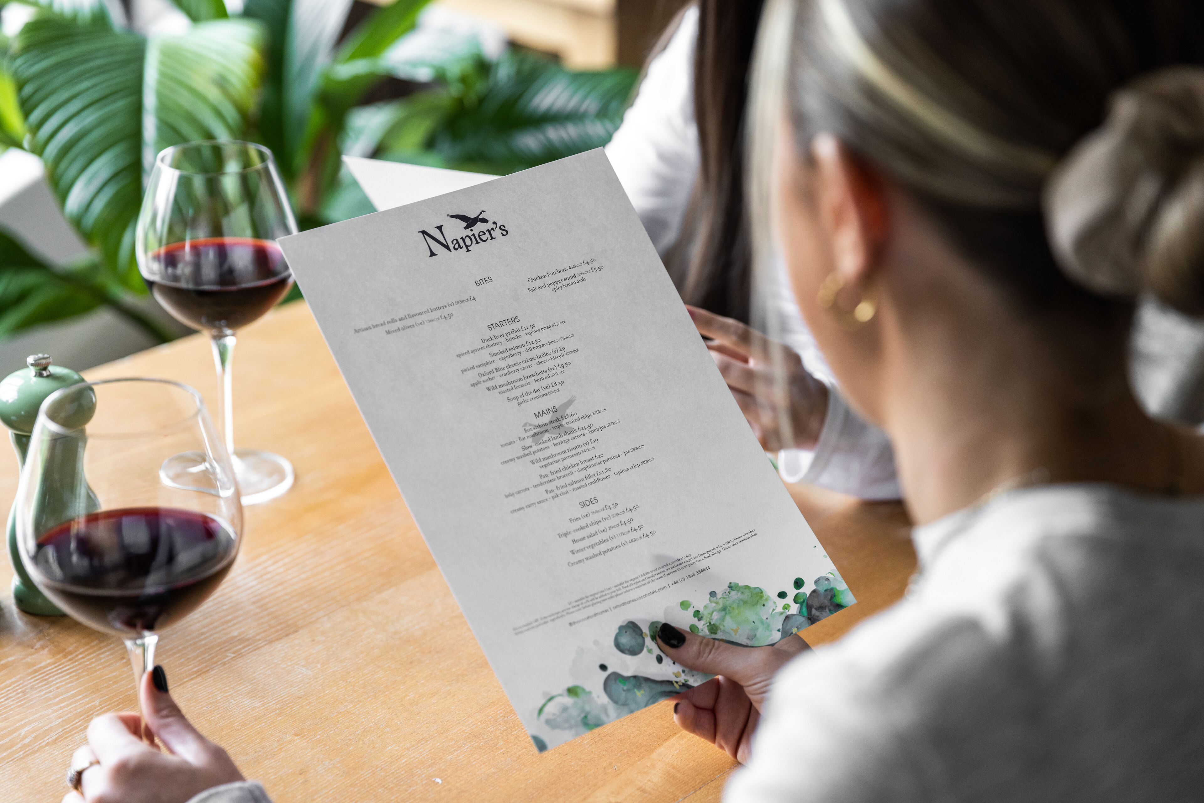 A person holding a menu at a restaurant with a wooden table and two glasses of red wine.