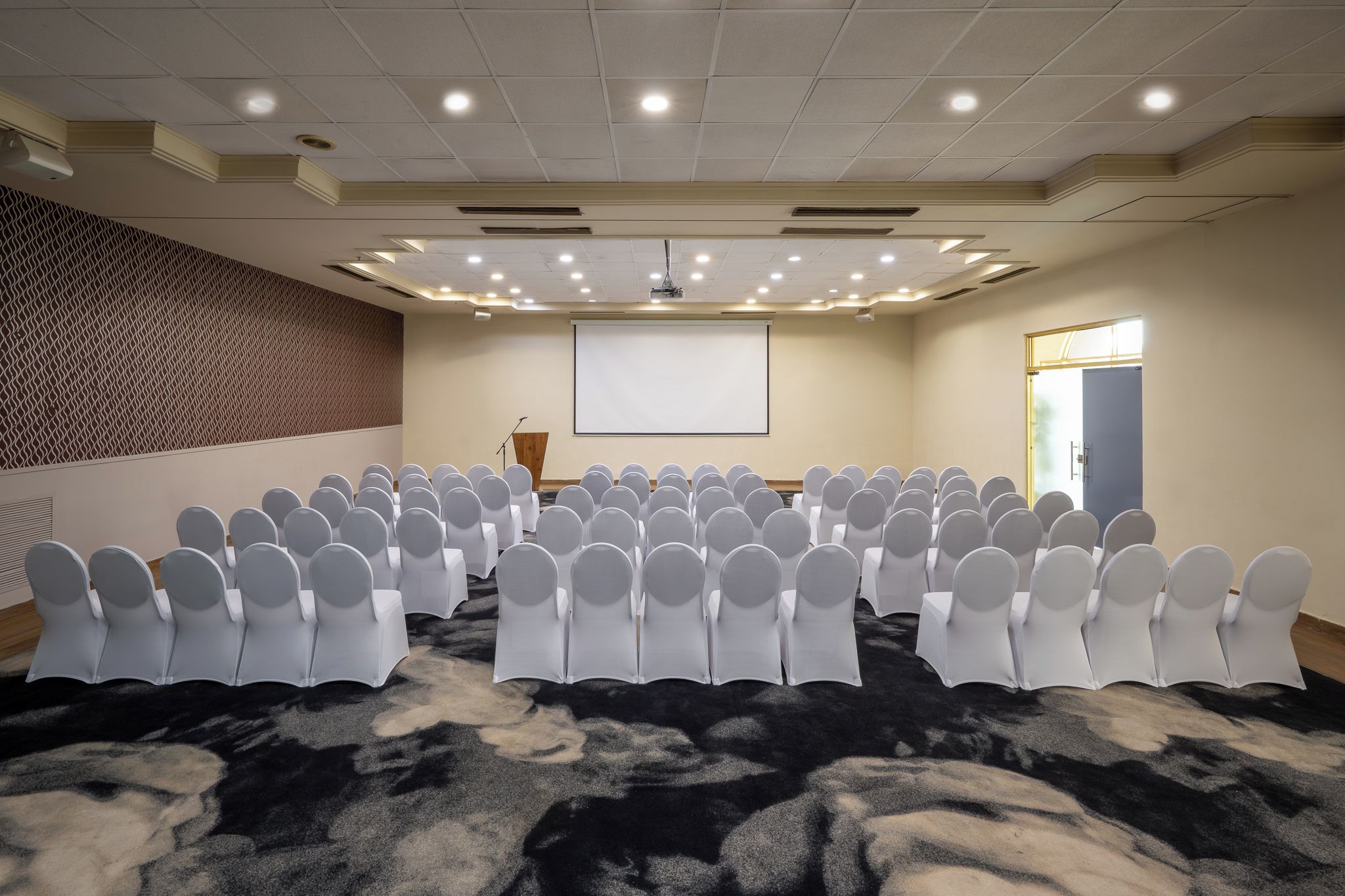 A conference room with rows of white chairs, a projector screen, and a carpeted floor.