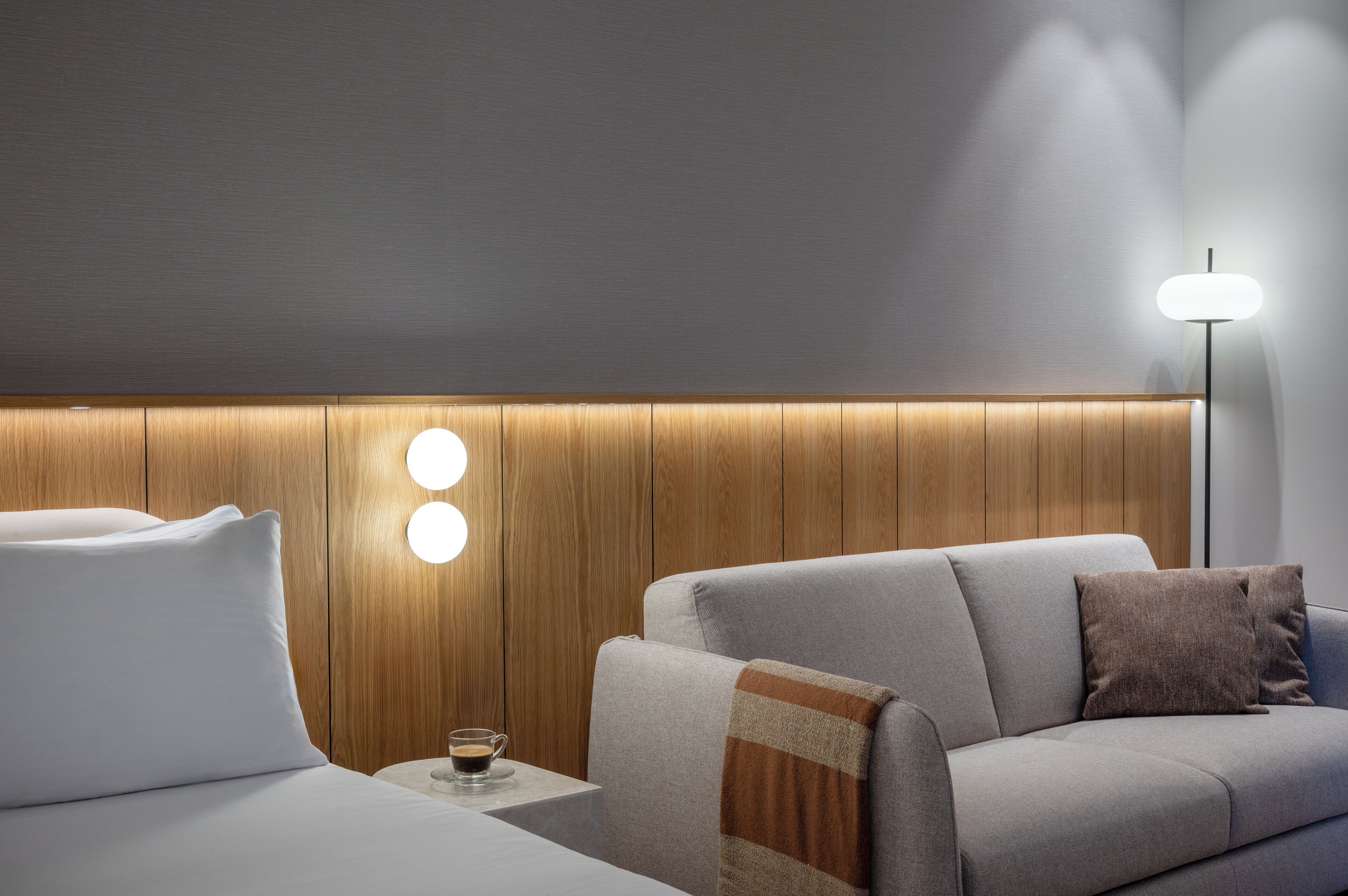 Una habitación de hotel moderna con un cabecero de madera, un sofá gris y una mesita de noche con una taza de café.