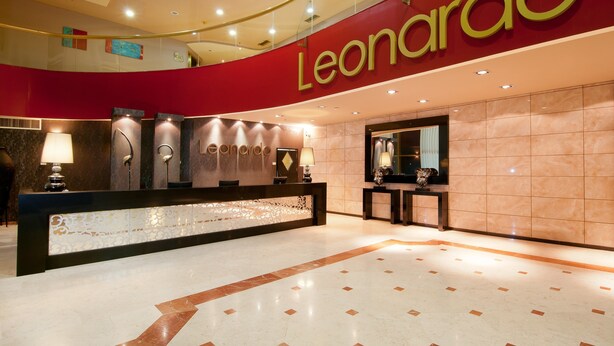 Zona de recepción de un hotel con un llamativo letrero 'Leonardo', un mostrador de recepción y un muro decorativo.