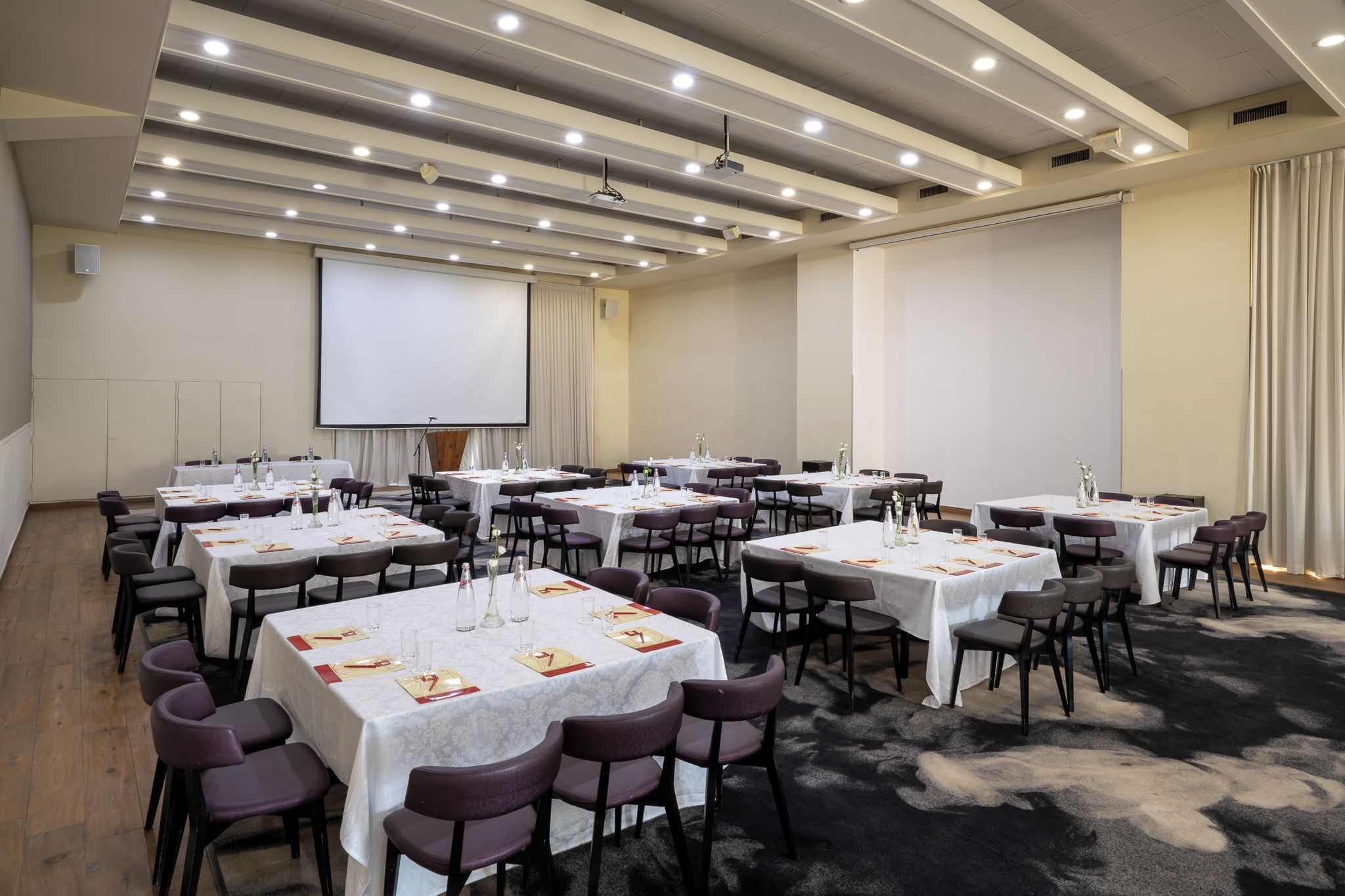Una sala de conferències amb diverses taules i cadires, una pantalla de projector i un terra amb moqueta.