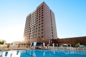 Un hotel alto con una zona de piscina en primer plano, que incluye tumbonas y sombrillas.