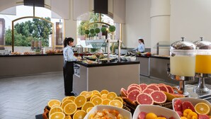 Zona de bufé con diversas frutas y bebidas, un empleado atendiendo el bufé, y vistas a árboles a través de grandes ventanales.