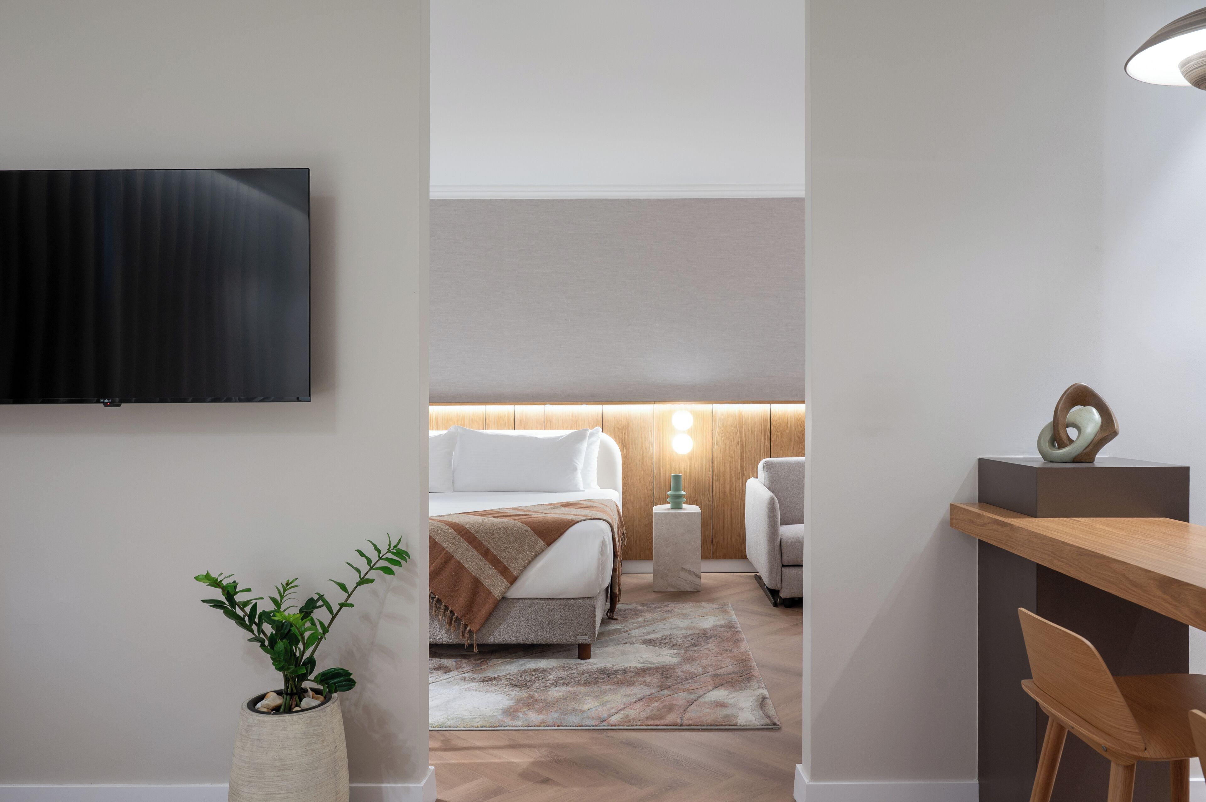 A modern hotel room with a bed, bedside table, lamp, and a flat-screen TV mounted on the wall.