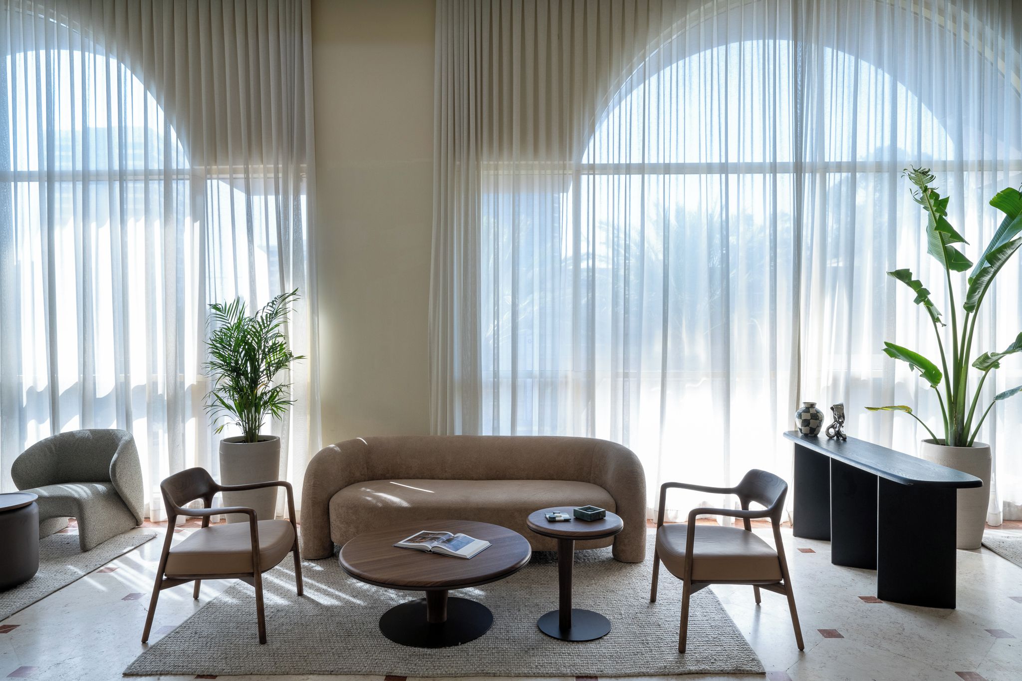 A modern living room with a beige sofa, two armchairs, a coffee table, and a side table. There are two large windows with sheer curtains, allowing natural light to fill the room.