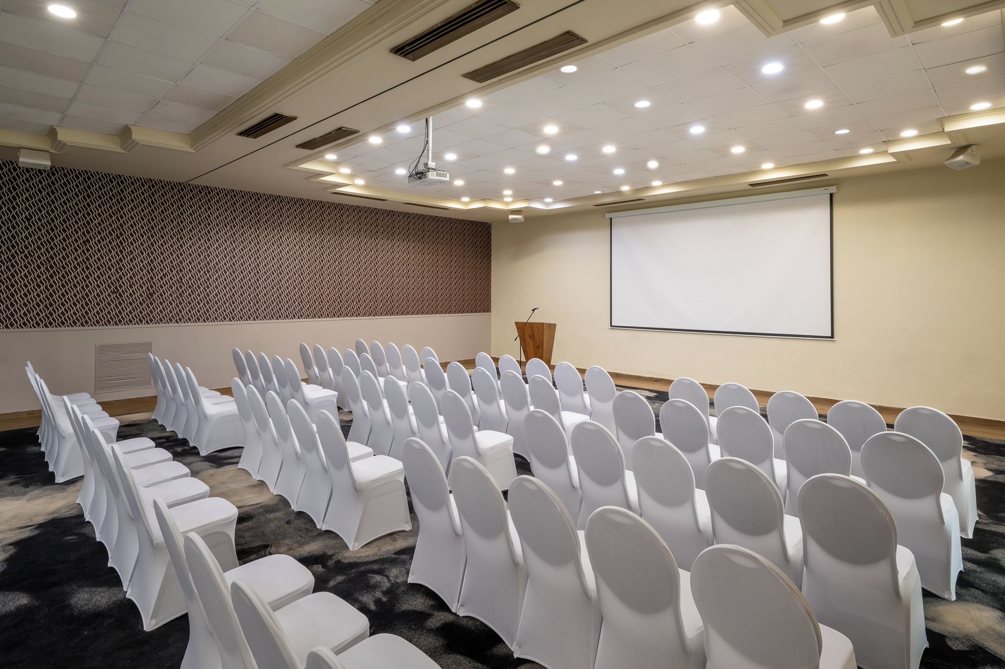 Une salle de conférence avec des rangées de chaises blanches, un pupitre, un écran de projection et un mur orné de motifs.