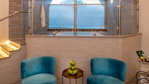 Un baño moderno con ducha de vidrio, tocador con bañera y vistas al exterior. Hay dos sillas azules y una pequeña mesa con manzanas.