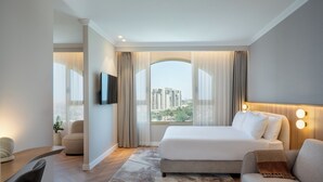 Una habitación de hotel moderna con una cama grande, un sofá, un televisor montado en la pared y vistas de la ciudad a través de un ventanal amplio.