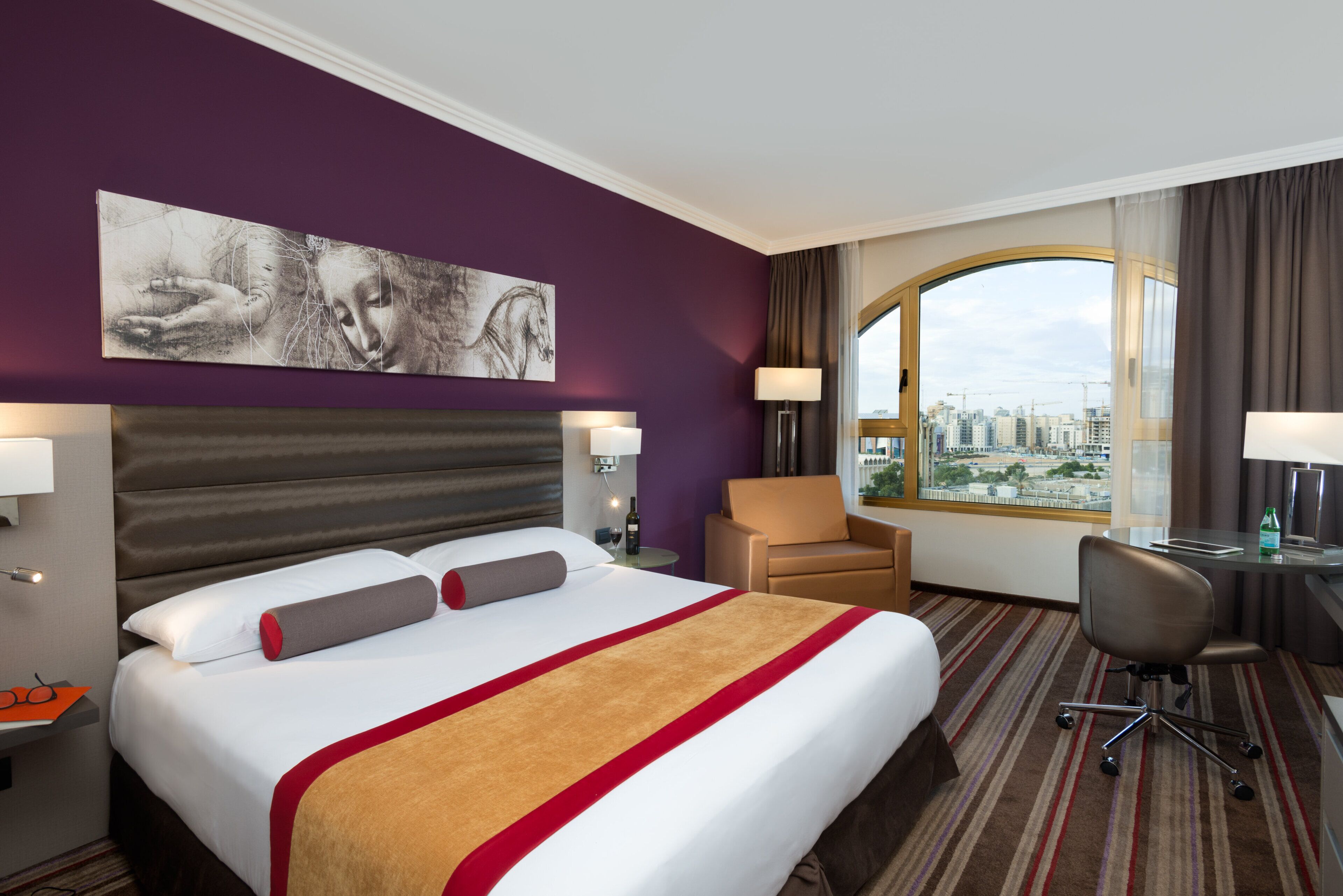 A hotel room with a large bed, a desk, a chair, and a view of the city.