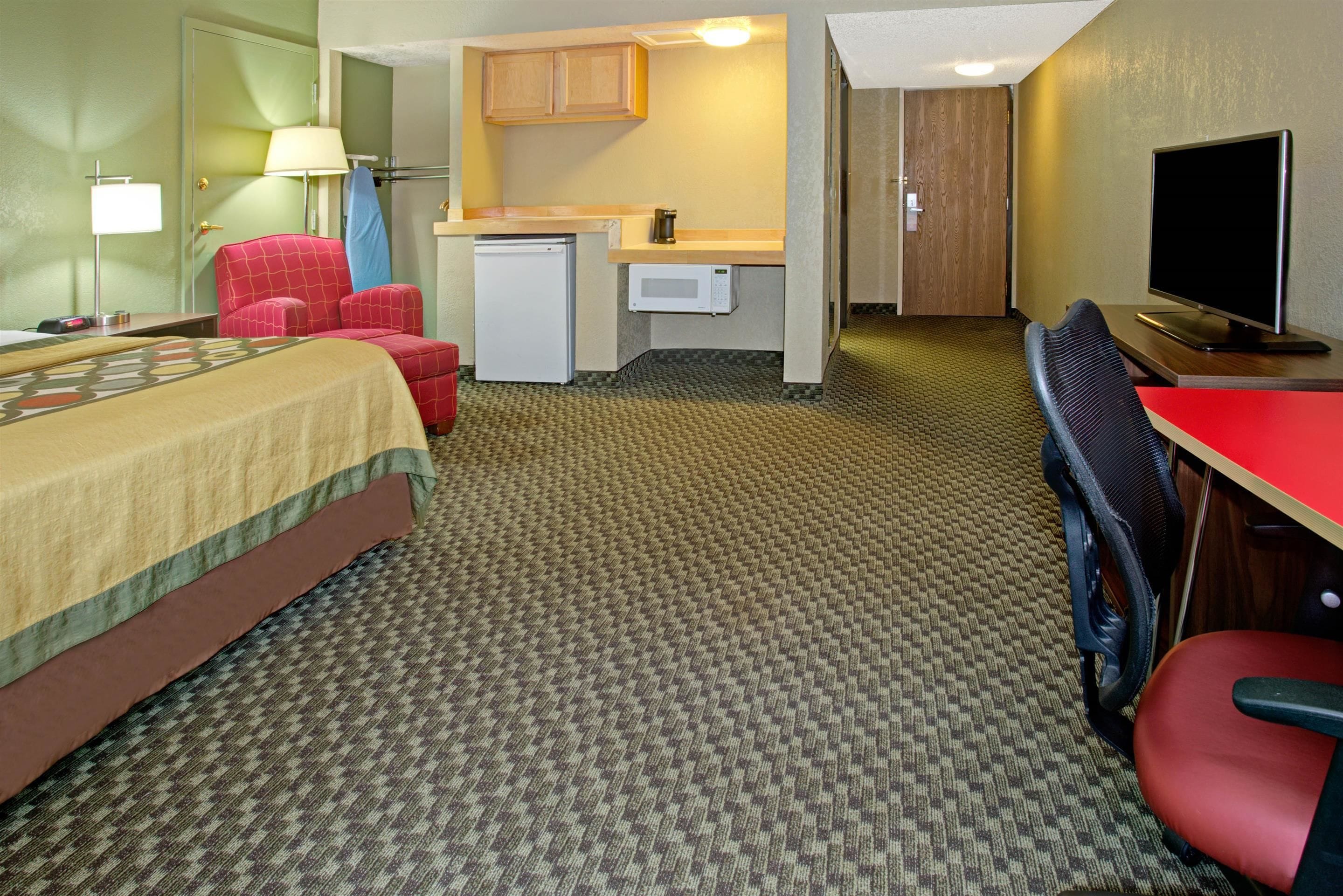 A hotel room with a bed, a desk with a computer, a microwave, a mini fridge, a red chair, and a television.