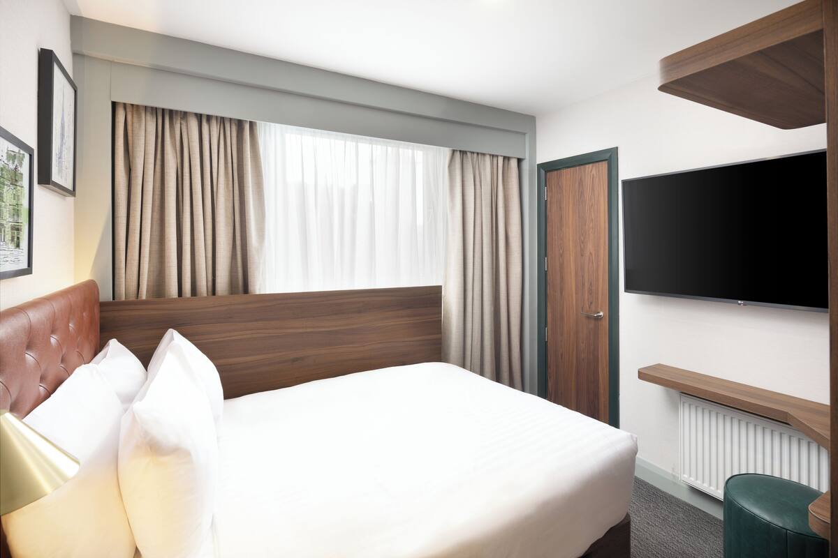 Habitación de hotel con una cama grande, televisión montada en la pared, cabecero de madera y una ventana con cortinas.