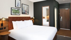 Habitación de hotel con una cama grande, cabecera de madera, una mesita redonda y una mesita de noche con lámpara. Se observan cuadros enmarcados en la pared y un espejo en la puerta.