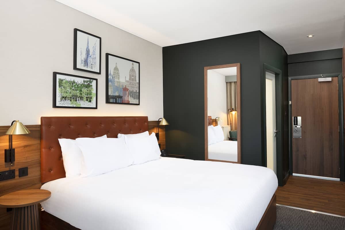 Habitación de hotel con una cama grande, cabecera de madera, una mesita redonda y una mesita de noche con lámpara. Se observan cuadros enmarcados en la pared y un espejo en la puerta.
