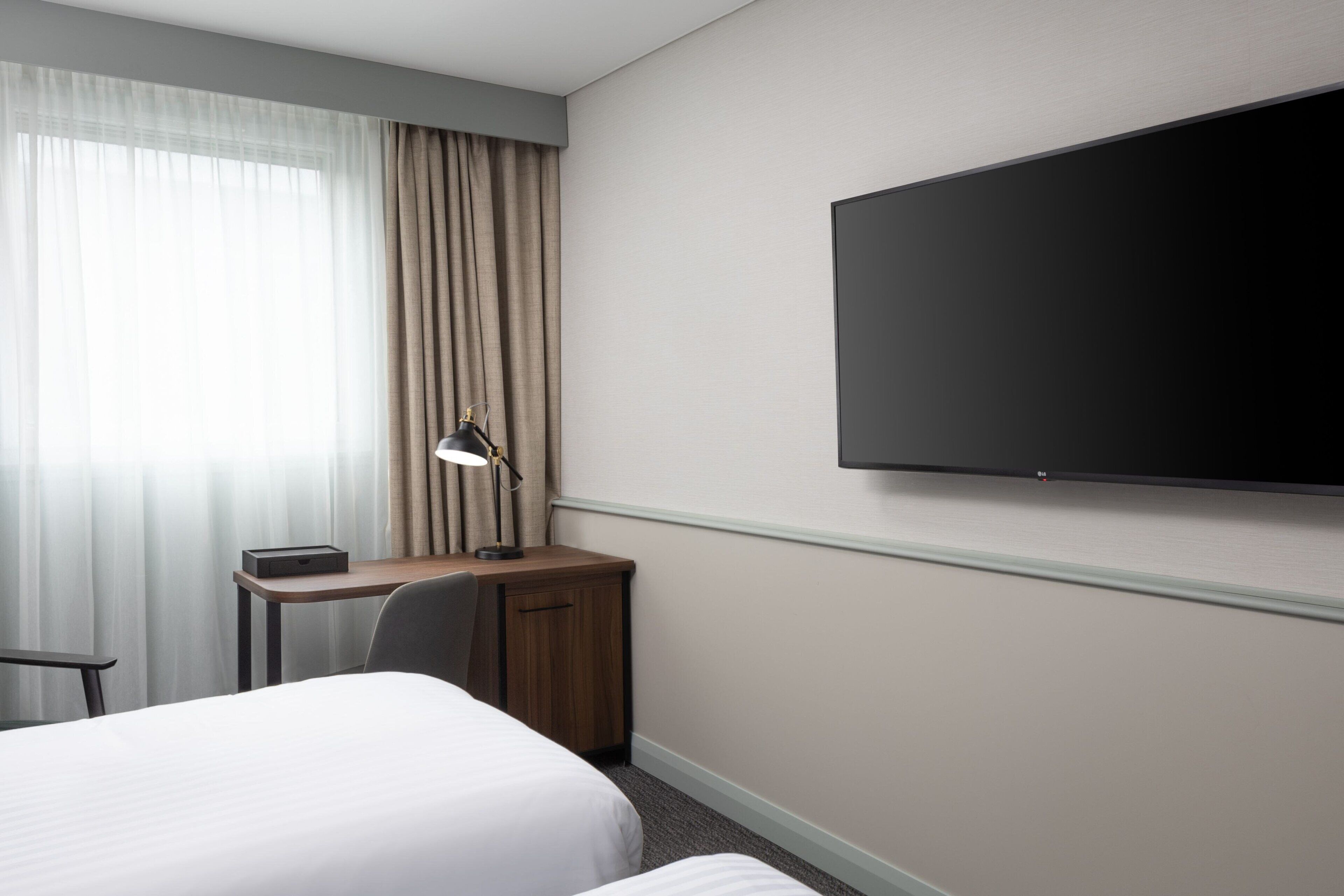 Une chambre d’hôtel avec deux lits, un bureau avec une lampe, un téléviseur à écran plat fixé au mur et une fenêtre avec des rideaux.