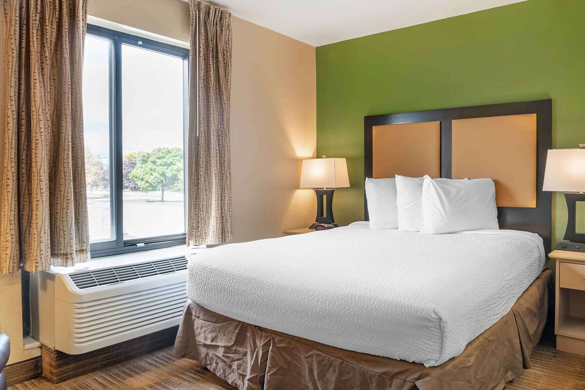 A hotel room with a large bed, two bedside lamps, a window with curtains, and a wall heater.
