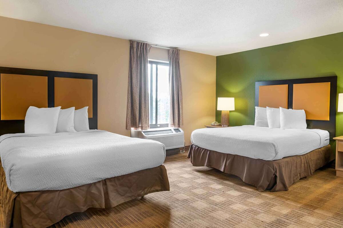 A hotel room with two beds, a window with curtains, an air conditioning unit, and a green accent wall.